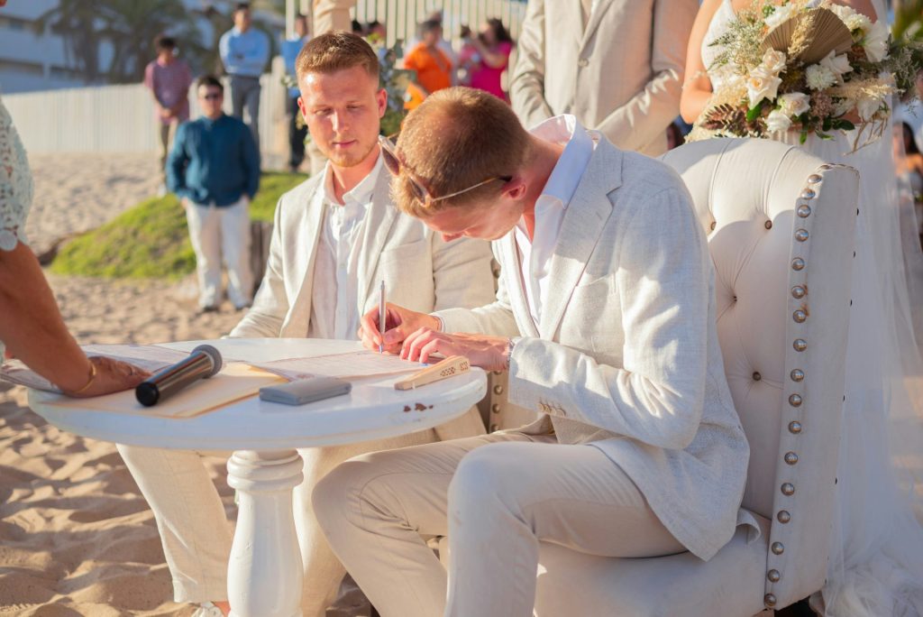 Two men in white suits signing wedding documents on beach